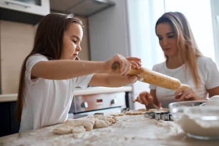 little child learning to work with rolling pin while mother controlling her, giving useful advice to daughter. close up side view photoの写真素材