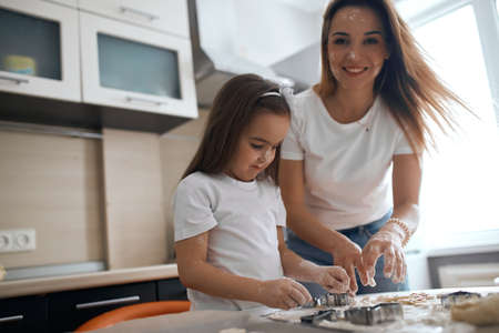 happy mother with flour on her face looking at the camera while her serious kid concentrated on...