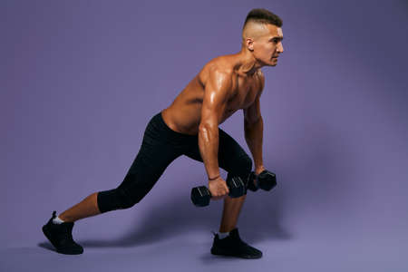young motivated atrong man doing loungers with dumbbells, full length side view photo, isolated blue...