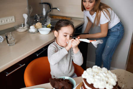 little hungry girl eating cream while cooking cake, mother wearing white T-shirt and jeans standing...