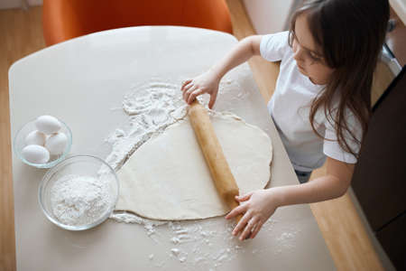 little girl in white T-shirt sitting at the kicthen table and rolling the dough, top view photoの写真素材