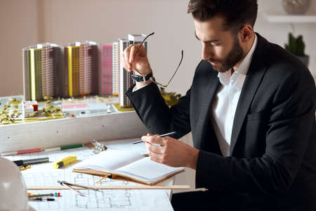 bearded good looking skilled architect making notes on notepad, close up side view photo.working...