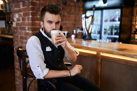 serious young man in elegant clothes drinking new sort of coffee, aroma which is not resisted by a...