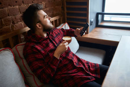 young thoughtful pensive man looking at the window, holding a smart phone and cup of tasty tea, thinking about business, life, womanの写真素材