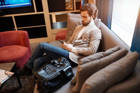 young attractive man in stylish casual clothes relaxing on the luxury comfortable sofa, using...