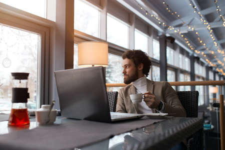 young serious man looking at the window, holding a cup, businessman having breakfast in the...