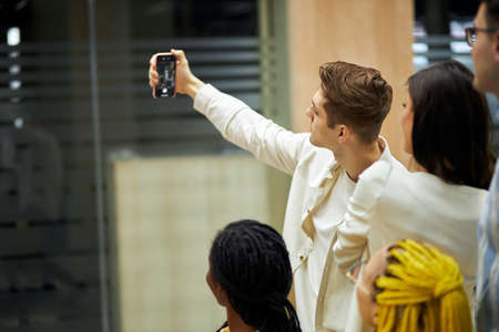handsome blonde guy holding his mpbile phone, taking selfiew with his colleagues, close up photo....