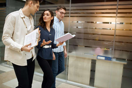 young beautiful brunette woman in elegant shirt giving business advice to men. young colleagues in smart casual wear discussing business while walking through the office corridor. copy space.の写真素材