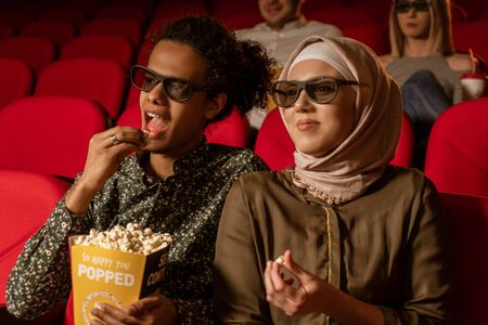 African american muslim man with his wife sitting in movie theater, watching 3D movie, eating...