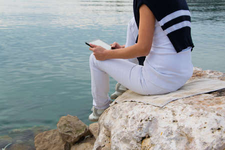 Crop view of attractive young woman  working with digital tablet sitting at seaside cliff edge....