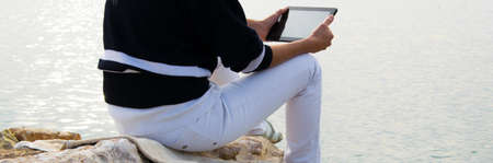 Crop view of attractive young woman  working with digital tablet sitting at seaside cliff edge....