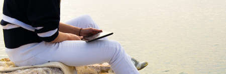 Crop view of attractive young woman  working with digital tablet sitting at seaside cliff edge....