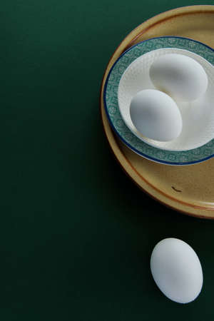Ceramic tablewares in warm pastel colors and fresh chicken eggs on deep green backgroundの写真素材