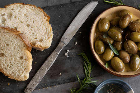 Italian sliced ciabatta bread on chopping board with herbs, extra virgin oil and marinated olives on dark grunge backdropの写真素材
