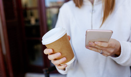 Close-up image of young hipster girl using smartphone at home interior, female hands typing on touch screen, social networking concept, coffee to go in hand.の写真素材