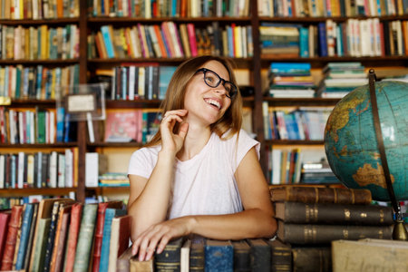 Young smiling attractive college girl portrait with eyeglasses standing in library, holding books....
