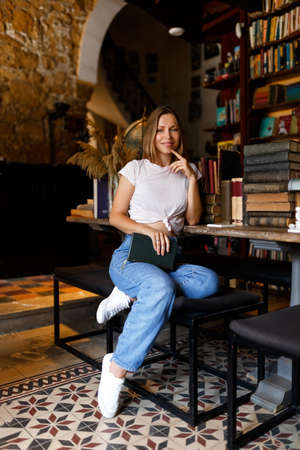Pretty smiling joyfully female with fair hair, dressed casually, looking with satisfaction at camera, being happy. Full body shot of good-looking beautiful woman sitting against bookshelf wallの写真素材