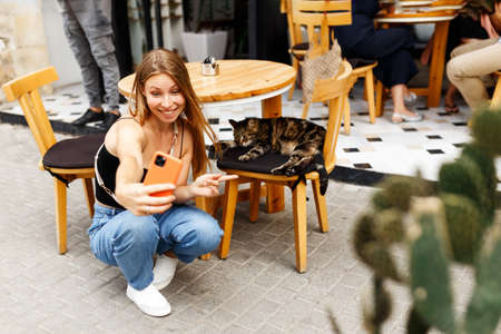 Portrait of attractive cheerful young girl using smartphone, blogger making selfie or live transmission with cat at downtown outdoors cafe placeの写真素材