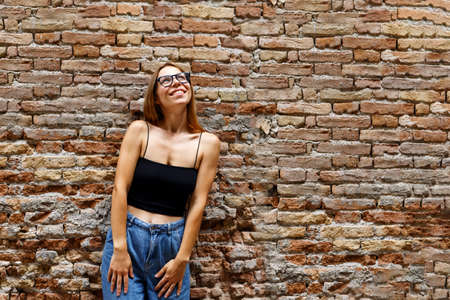 A hipster girl with long brown hair wearing a denim is looking aside while standing on a brick wall background on a street. Empty space for text o designの写真素材