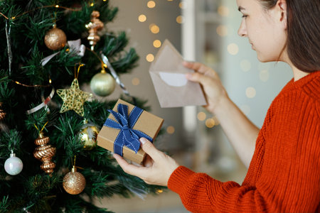 Woman opening gift box next to xmas tree and reading greeting card. Christmas time, New Year...