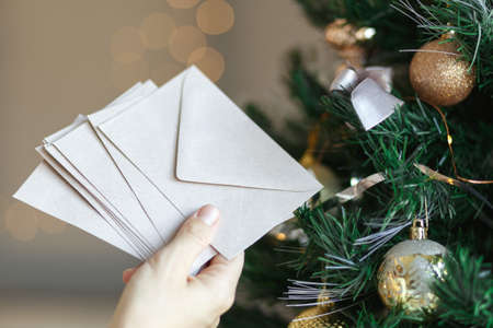 Hand holding stack of craft paper envelopes for christmas greeting card invitation design on christmas tree background with bokehの写真素材