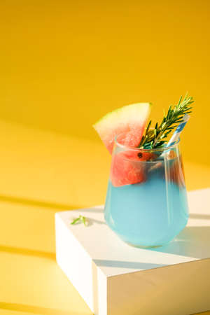 Blue margarita alcoholic cocktail with tequila on pedestal, liqueur,watermelon and ice, summer yellow background, copy space, shadowsの写真素材