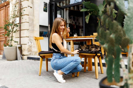 A young hipster woman sits at a table at an outdoor cafe bar and plays with the cat. Summertimeの写真素材