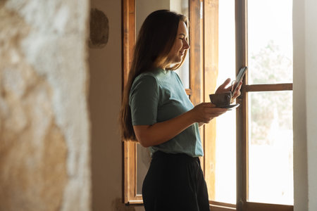 Profile view of beautiful woman drinking first morning coffee by the window and reading news and...