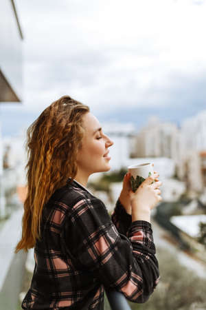 A young, brunette woman dressed in sleepwear enjoys her morning coffee outdoors on the terrace under warm sunlight. City lifestyleの写真素材