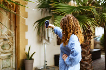 Unrecognizable woman in blue sweater with brunette hair streaming with smart phone in hand, shooting social media blog in modern outdoor cafe.Hipster Influencer using social networksの写真素材