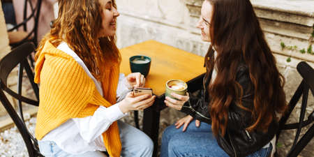 Cheerful female best friend share multimedia files on smartphones spending free time together,smiling hipster girls drinking coffee, sitting at terrace cafeの写真素材
