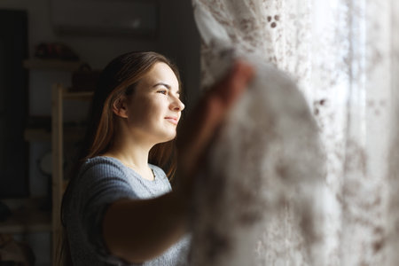 Pretty young woman in apartment opening window curtains after wake up, enjoying morning...