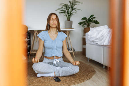 Trendy young woman wearing earphones meditating online and listening music on smartphone player app while sitting on floor at home,meditation app as part of mindfulness morning routine,peaceful moodの写真素材