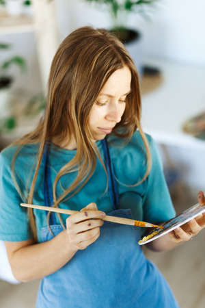 Woman artist holding paintbrush.Creative woman enjoying painting.Young beautiful woman in art studio, taking colorful paints from palette,creating masterpiece on easel, work, imagination.の写真素材