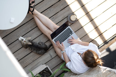 Top-down of young female copywriter typing text on laptop keyboard sitting on wooden terrace with...