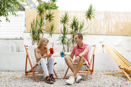 Happy young couple with glasses of lemonade sitting on deck chairs at sea beach or cafe placeの写真素材