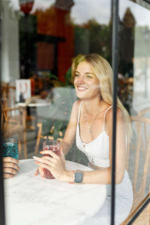 Young blonde happy woman drinking lemonade in the cafe, view through windowの写真素材