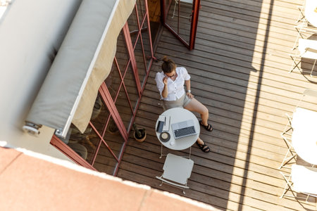 Top-down of young female copywriter typing text on laptop keyboard sitting on wooden terrace with...