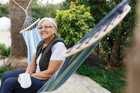 Beautiful senior blonde woman reading book and sitting in hammock in the gardenの写真素材