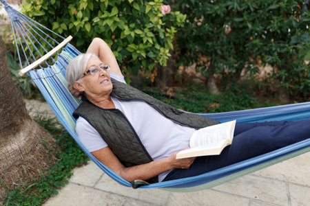 Smiling senior good-looking grey hair woman wearing glasses while reading in hammock in the summer gardenの写真素材