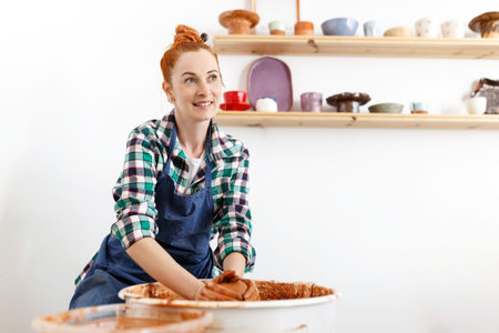 Cheerful female sculptor while she making clay pot on pottery wheel. Idea of small business and...