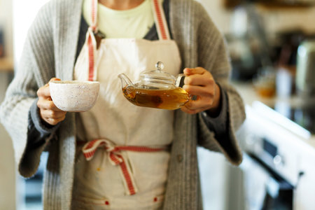Cropped view of young woman spending morning standing on kitchen, pouring traditional tea from transparent teapot into a cup. Female make hot drink with organic herbs.の写真素材