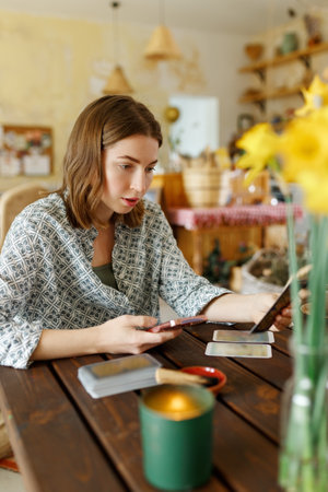 Beautiful young woman is guessing on cards with tarot, runes on wooden table and uses an online app in phone to interpret divination, home interior,candles and dry herbsの写真素材