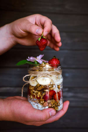 Granola in a glass jar with strawberries, bananas, yogurt and guns in hands on a black wooden backgroundの写真素材
