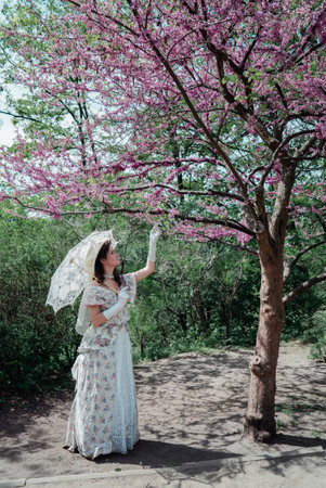 girl bride in vintage dress with white lace umbrella in park near blossoming tree with pink flowersの写真素材