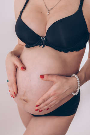 Pregnant woman with red nails manicure in cool black underwear holding her belly on white background, close upの写真素材