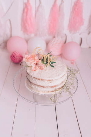 One year birthday party white cake with rose flower and golden letters. Pink balloons on white wooden backgroundの写真素材