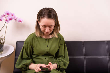 Young beautiful woman in gorgeous green dress sitting in queue on black leather sofa with phone in handsの写真素材