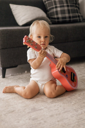 Cute blonde little baby playing on a childrens pink guitar sitting on the floor at homeの写真素材