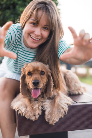 Young beautiful woman sitting on a bench and taking selfie with her dog. playing and having fun with her cocker spaniel dog, summer sunset in public parkの写真素材
